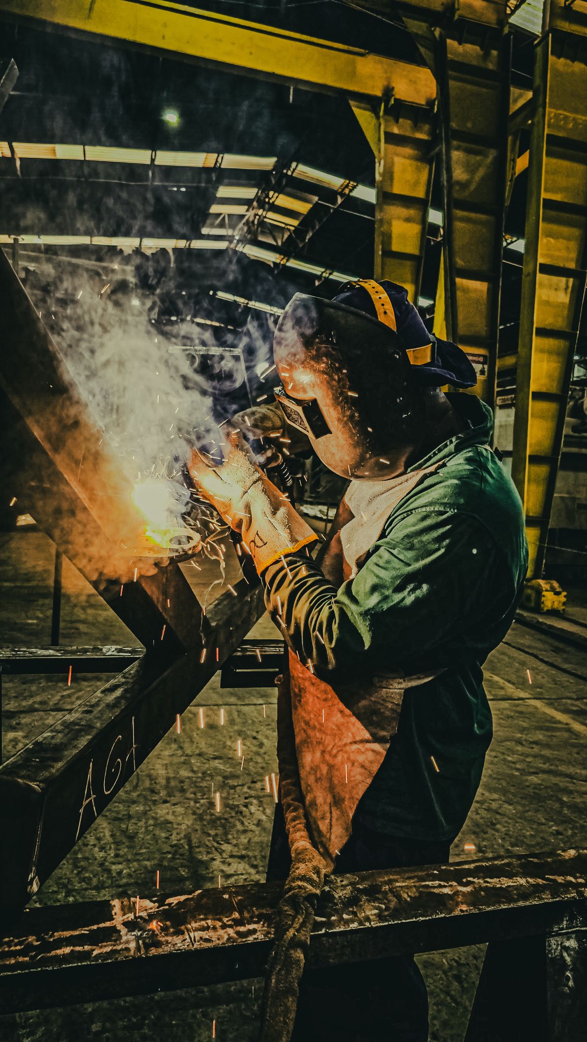 Soldador com EPI completo montando estrutura metálica em galpão industrial.